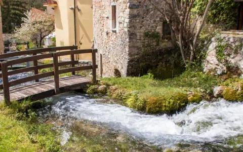 Un ponticello di legno attraversa un ruscello gorgogliante, con antiche costruzioni in pietra e un ciliegio in fiore. Un angolo di pace in Umbria dove andare.