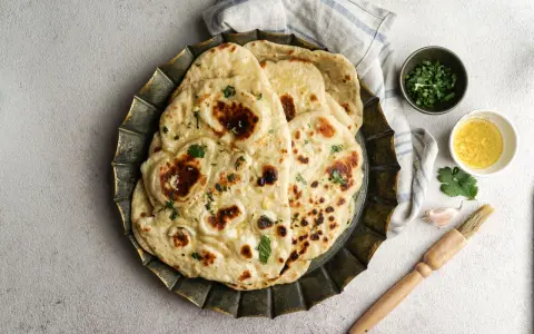 Pane in padella soffice e dorato, guarnito con erbe fresche. Accanto, ciotoline con olio e coriandolo, uno spicchio d'aglio e un pennello da cucina.