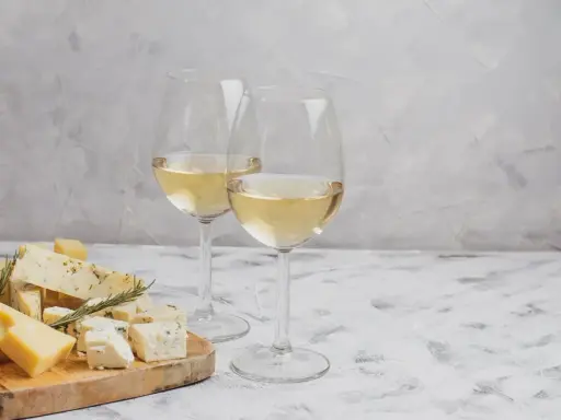 Due calici di Sauvignon Gris dorato accompagnano un tagliere di formaggi misti, tra cui erborinato e stagionato, su un tavolo grigio.
