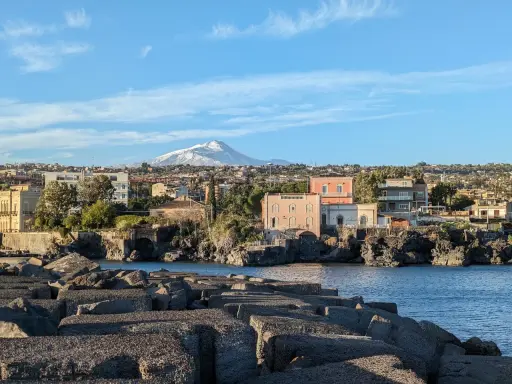Vista sul mare con blocchi di cemento in primo piano e un panorama di case con l'Etna innevato sullo sfondo, perfetto per un itinerario a Catania.