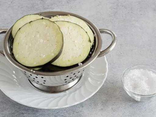 Fette di melanzane salate in un colino, pronte per spurgare. Accanto, una ciotolina di sale grosso.