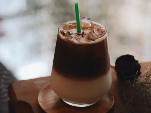 Un bicchiere di **caffè freddo** stratificato con ghiaccio e una cannuccia verde, su un tavolino di legno.