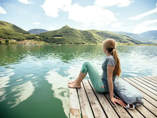 Ragazza con zaino seduta su un pontile di legno, ammira il **lago di Caldaro** e le montagne verdi sotto un cielo azzurro con nuvole bianche.
