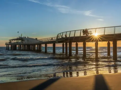 Un molo moderno si estende sul mare al tramonto, con la costa toscana mappa sullo sfondo. Le onde lambiscono la spiaggia dorata.