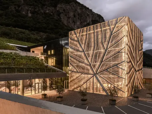 Cantina Sociale Bolzano: edificio moderno con facciata luminosa a forma di foglia, circondato da vigneti e montagne.