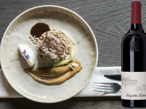 Piatto gourmet con carne, cereali e salsa, accanto a una bottiglia di Lagrein Riserva. Perfetti lagrein abbinamenti per un'esperienza culinaria raffinata.
