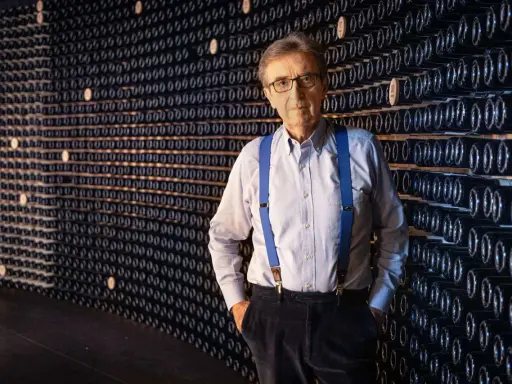 L'enologo Cotarella, con le sue bretelle blu, posa con sicurezza davanti a una parete di bottiglie di vino.