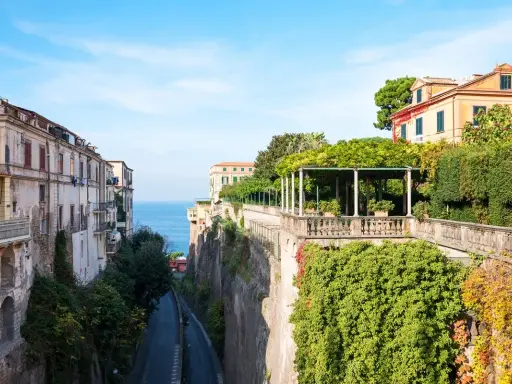 Edifici affacciati sul mare blu, con un viale alberato che scende verso la costa. La **penisola sorrentina** offre scorci mozzafiato tra ville storiche e vegetazione rigogliosa.