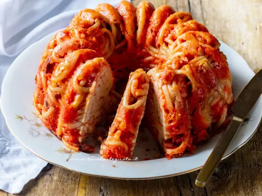 Un invitante timballo di pasta, con spaghetti avvolti in un ricco sugo rosso, servito su un piatto bianco con un coltello dorato.