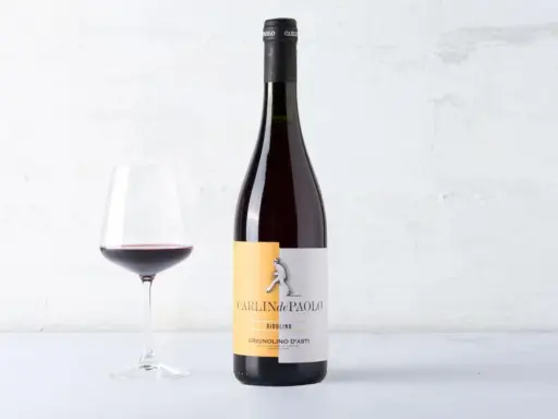 Calice di vino rosso accanto a una bottiglia di Grignolino d'Asti Carlin de Paolo. Un vino che ricorda il barbera vitigno per la sua vivacità.