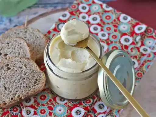 Un cucchiaio dorato preleva una generosa porzione di maionese con uovo sodo da un barattolo di vetro. Fette di pane rustico completano la scena.