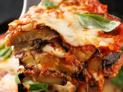 Una fetta di parmigiana di melanzane non fritte, con strati di melanzane, sugo e mozzarella filante, guarnita con basilico fresco.