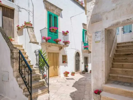 Un vicolo fiorito a Locorotondo, con scalinate in pietra bianca, balconi verdi e gerani rosa.
