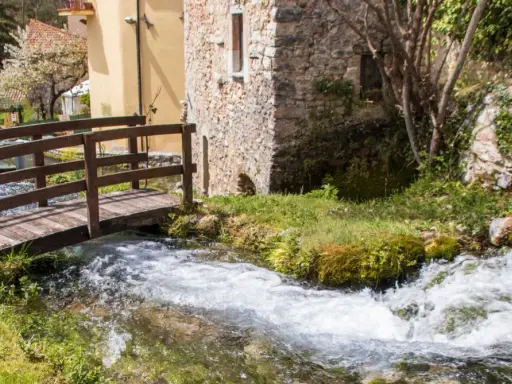 Un ponticello di legno attraversa un ruscello gorgogliante, con antiche costruzioni in pietra e un ciliegio in fiore. Un angolo di pace in Umbria dove andare.