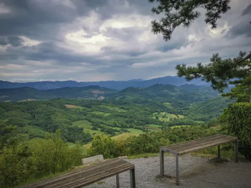 Panoramica del **Carso** con colline verdi e montagne all'orizzonte, sotto un cielo nuvoloso. Due panchine invitano alla sosta.