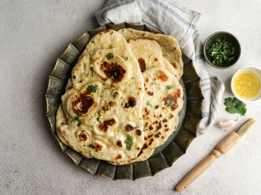 Pane in padella soffice e dorato, guarnito con erbe fresche. Accanto, ciotoline con olio e coriandolo, uno spicchio d'aglio e un pennello da cucina.