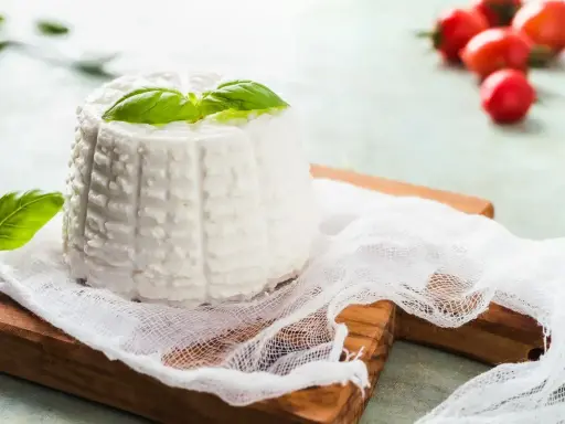 Ricotta fatta in casa, fresca e profumata, guarnita con basilico su un tagliere di legno. Sullo sfondo, pomodorini ciliegino.