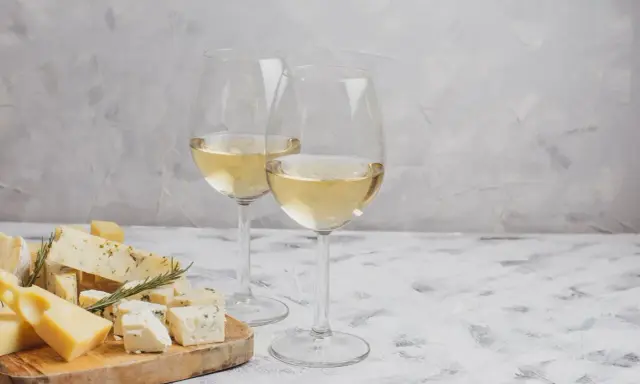 Due calici di Sauvignon Gris dorato accompagnano un tagliere di formaggi misti, tra cui erborinato e stagionato, su un tavolo grigio.