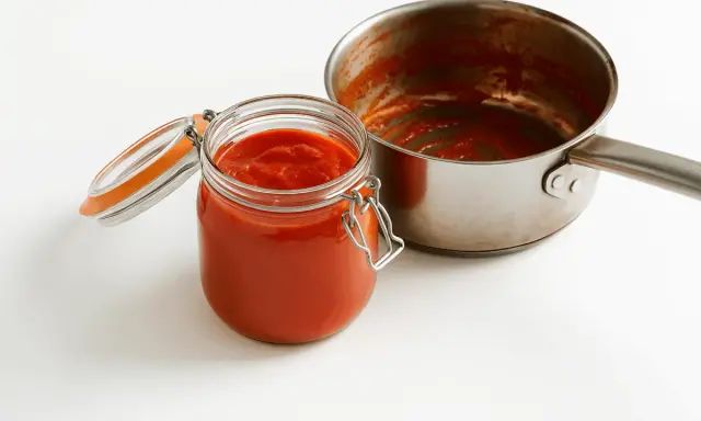 Barattolo di vetro pieno di conserva di pomodoro, accanto a una pentola con residui rossi.