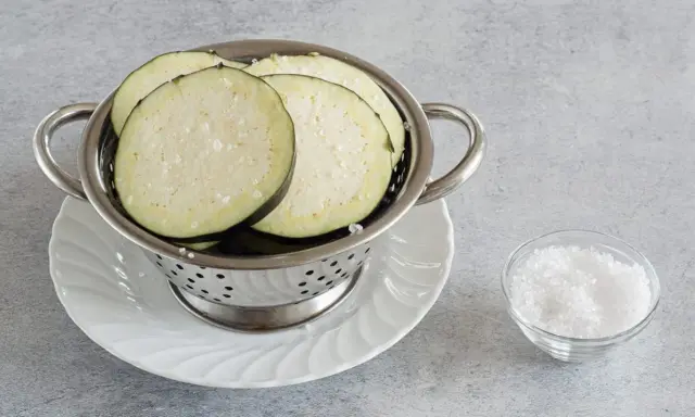 Fette di melanzane salate in un colino, pronte per spurgare. Accanto, una ciotolina di sale grosso.