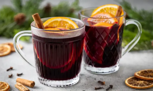 Due tazze fumanti di vin brulé, decorate con arancia e cannella, su un tavolo con spezie e rami di pino.