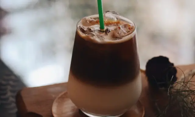 Un bicchiere di **caffè freddo** stratificato con ghiaccio e una cannuccia verde, su un tavolino di legno.