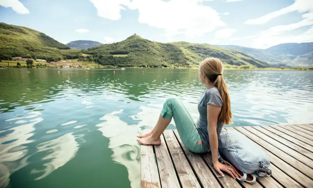 Ragazza con zaino seduta su un pontile di legno, ammira il **lago di Caldaro** e le montagne verdi sotto un cielo azzurro con nuvole bianche.