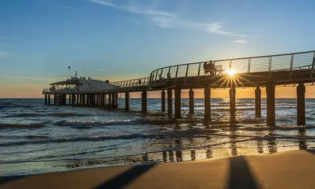 Un molo moderno si estende sul mare al tramonto, con la costa toscana mappa sullo sfondo. Le onde lambiscono la spiaggia dorata.