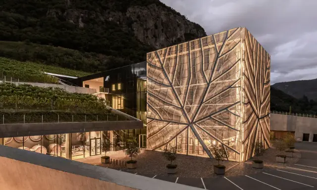 Cantina Sociale Bolzano: edificio moderno con facciata luminosa a forma di foglia, circondato da vigneti e montagne.