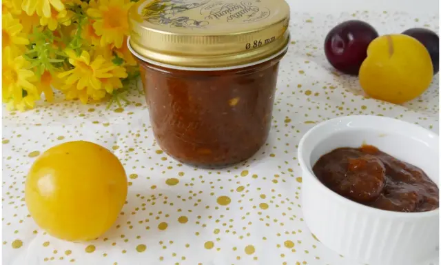Vasetto di marmellata di susine fatto in casa, con prugne rosse e gialle e fiori gialli sullo sfondo.