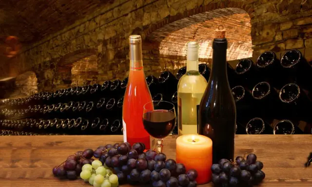 Uva e bottiglie di vino in una cantina storica, con un bicchiere di rosso e una candela. I vitigni Franciacorta sono pronti per essere degustati.