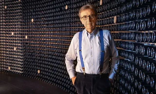 L'enologo Cotarella, con le sue bretelle blu, posa con sicurezza davanti a una parete di bottiglie di vino.