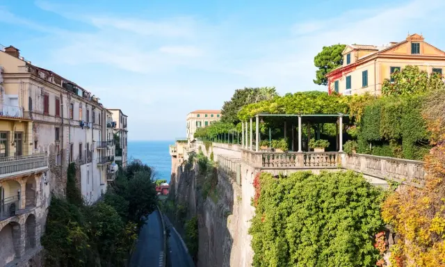 Edifici affacciati sul mare blu, con un viale alberato che scende verso la costa. La **penisola sorrentina** offre scorci mozzafiato tra ville storiche e vegetazione rigogliosa.