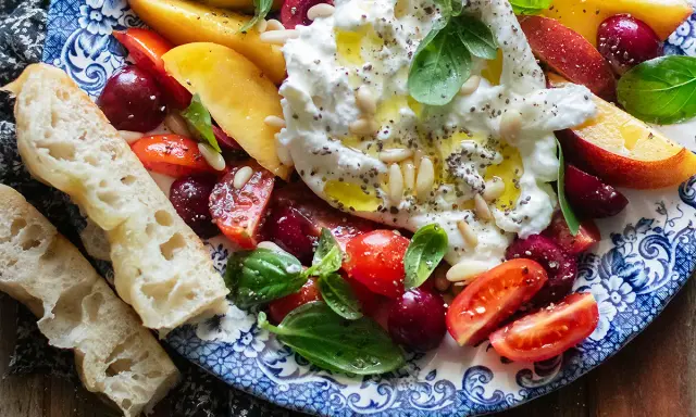 Insalata estiva con burrata, pesche, ciliegie, pomodorini e basilico, condita con olio e pinoli. Accompagnata da pane casereccio.