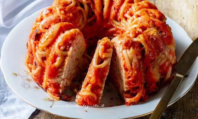 Un invitante timballo di pasta, con spaghetti avvolti in un ricco sugo rosso, servito su un piatto bianco con un coltello dorato.