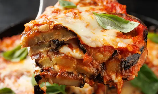 Una fetta di parmigiana di melanzane non fritte, con strati di melanzane, sugo e mozzarella filante, guarnita con basilico fresco.