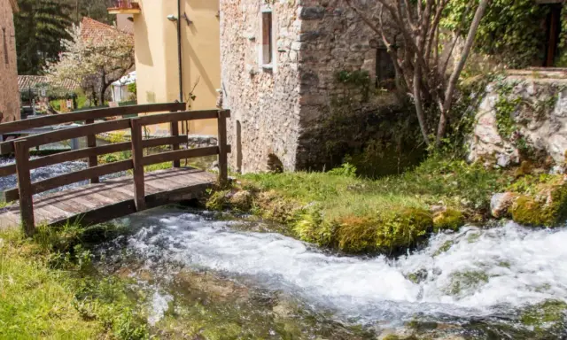 Un ponticello di legno attraversa un ruscello gorgogliante, con antiche costruzioni in pietra e un ciliegio in fiore. Un angolo di pace in Umbria dove andare.