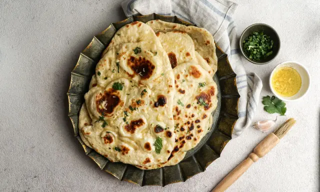 Pane in padella soffice e dorato, guarnito con erbe fresche. Accanto, ciotoline con olio e coriandolo, uno spicchio d'aglio e un pennello da cucina.