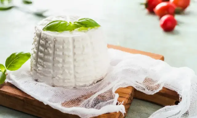 Ricotta fatta in casa, fresca e profumata, guarnita con basilico su un tagliere di legno. Sullo sfondo, pomodorini ciliegino.