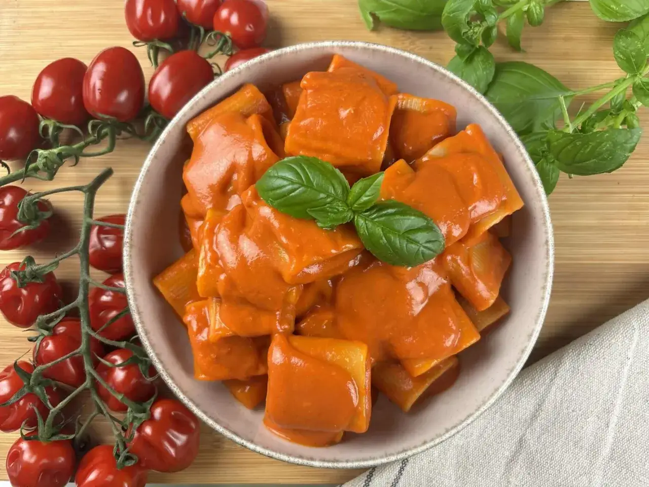 Un piatto di paccheri alla Vittorio, conditi con una cremosa salsa al pomodoro e guarniti con basilico fresco. Accanto, pomodorini maturi e foglie di basilico.