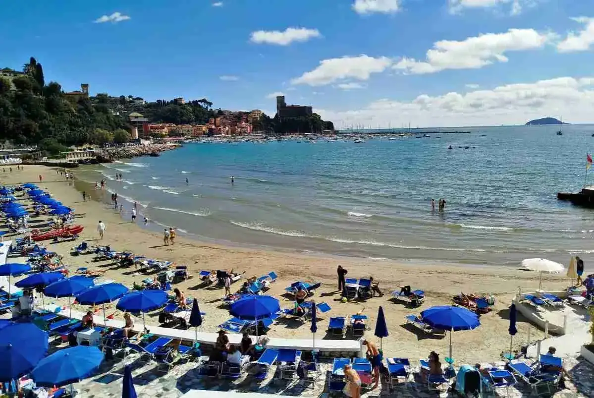 Spiaggia affollata con ombrelloni blu e lettini, mare azzurro e castello sullo sfondo. Una cartina della Liguria Levante ideale per le vacanze.