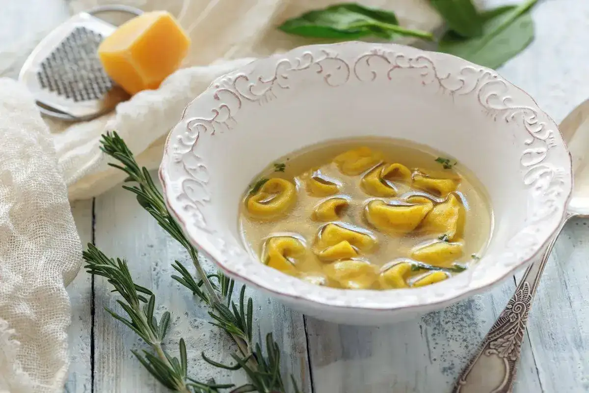 Un invitante piatto di tortellini in brodo, con rosmarino fresco e un pezzo di formaggio grattugiato sullo sfondo.