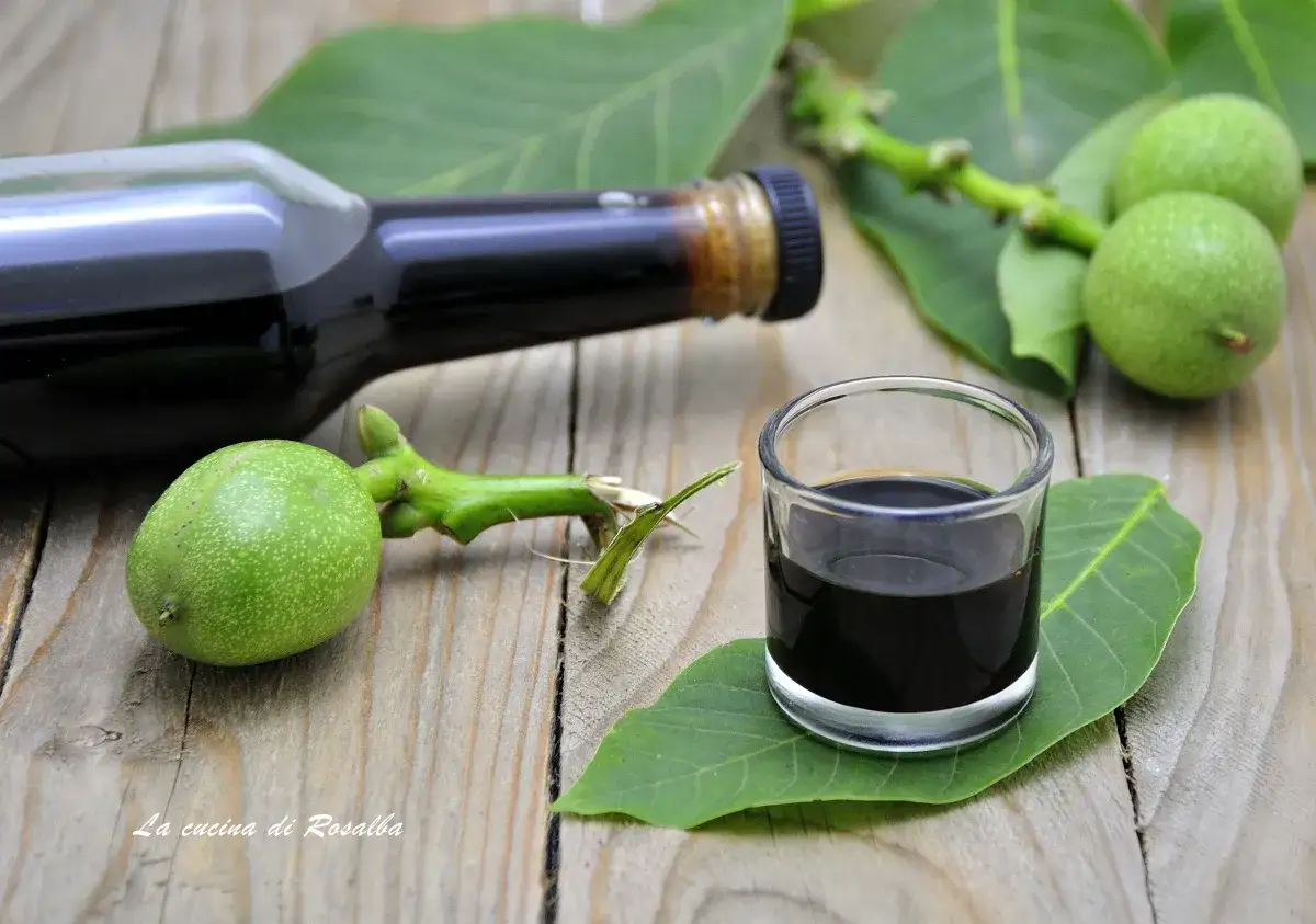 Un bicchierino di nocino scuro, con noci verdi fresche e foglie su un tavolo di legno. La ricetta del nocino &egrave; pronta per essere gustata.