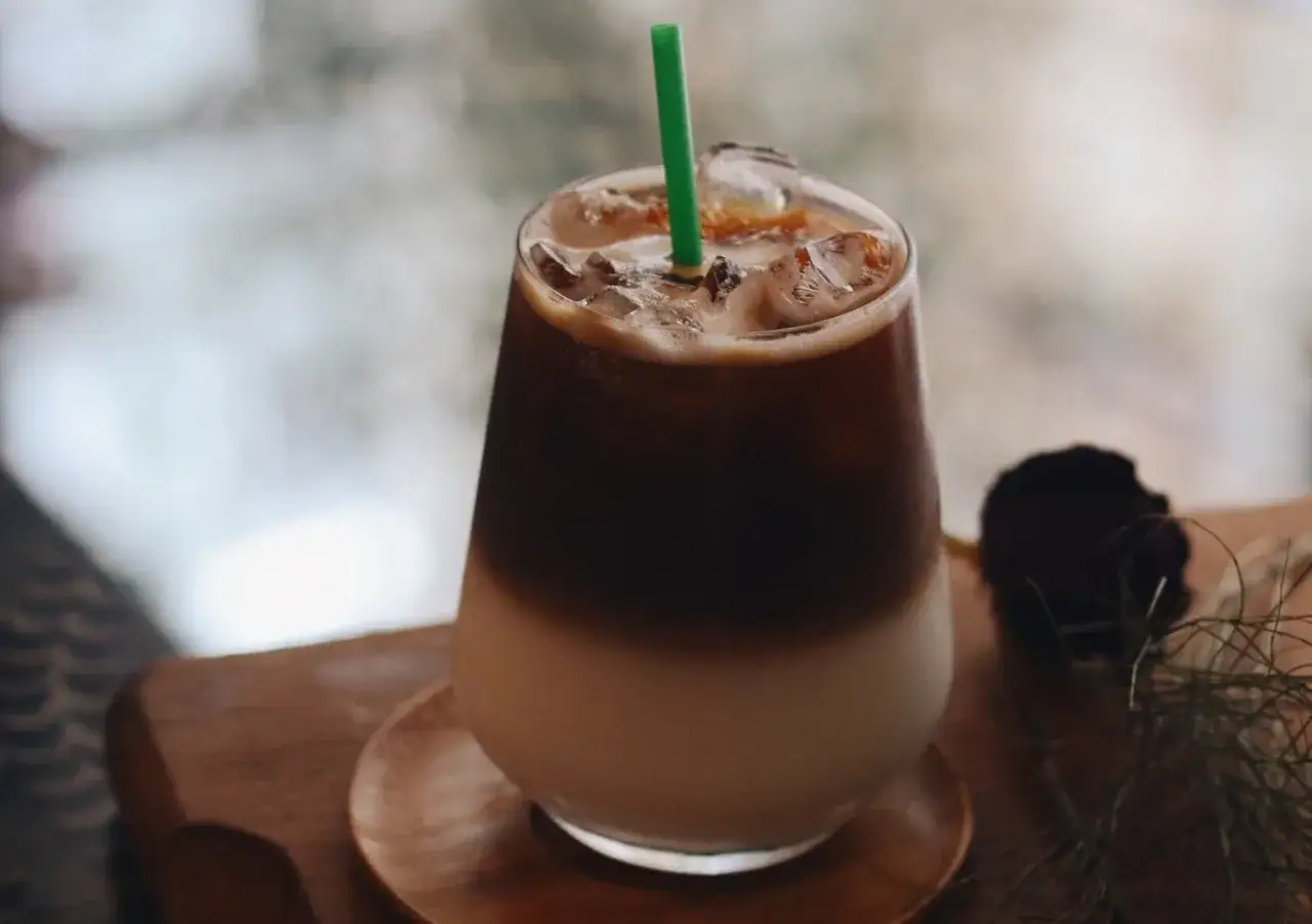 Un bicchiere di caff&egrave; freddo stratificato, con ghiaccio e una cannuccia verde, su un tavolino di legno.