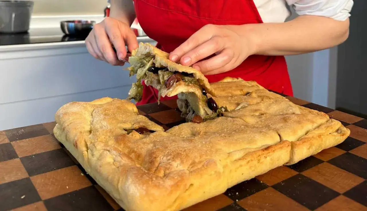 Una fetta generosa di pizza di scarola, ripiena di verdure e olive, viene sollevata da una teglia dorata.