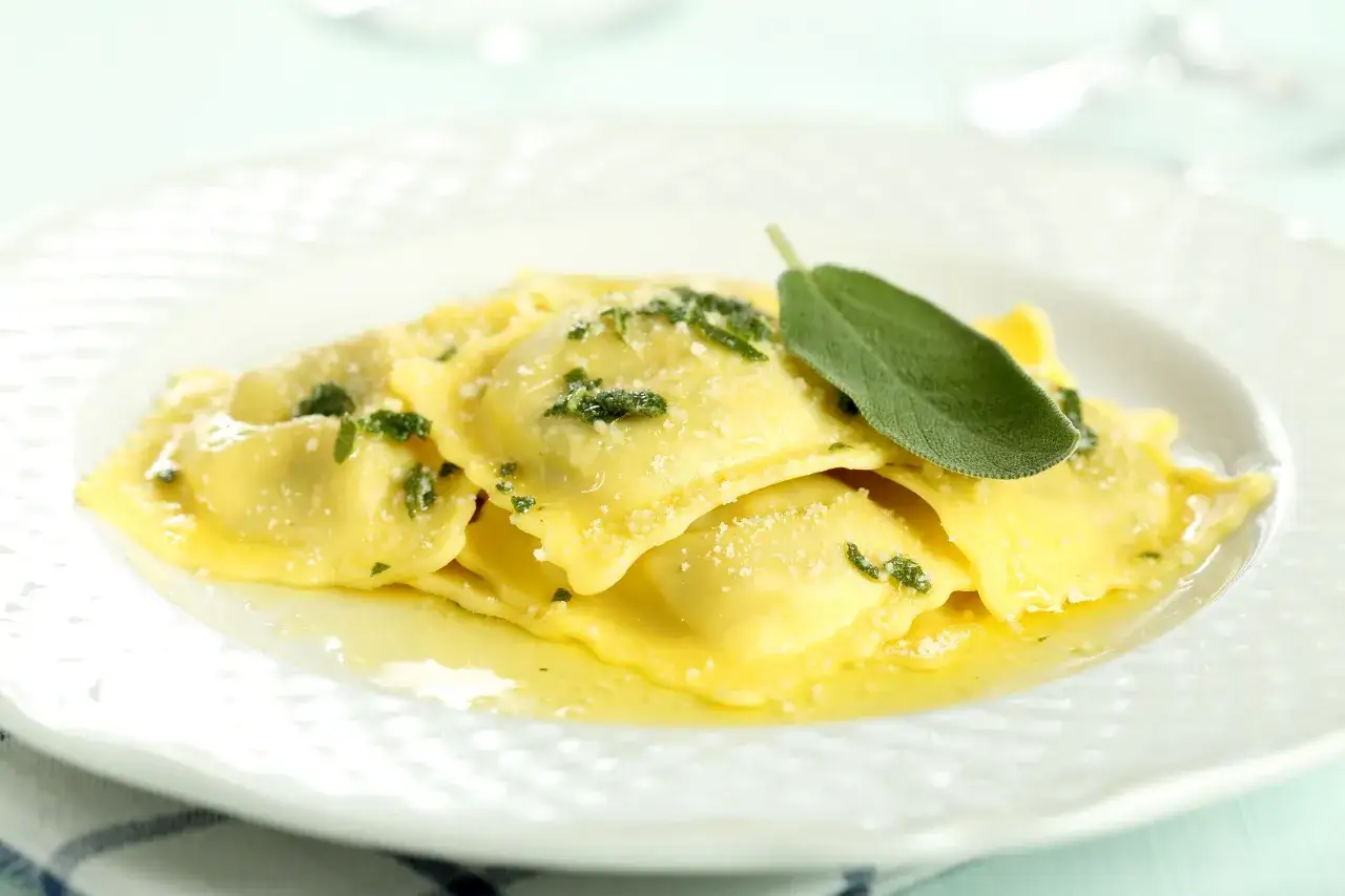 Ravioli dorati conditi con burro fuso, parmigiano e un tocco aromatico di pesto di salvia, guarniti con una foglia fresca.
