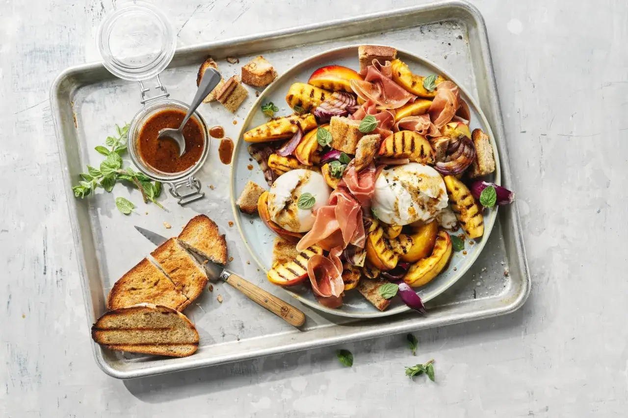 Insalata estiva con burrata, pesche grigliate, prosciutto crudo e crostini. Un piatto fresco e saporito.