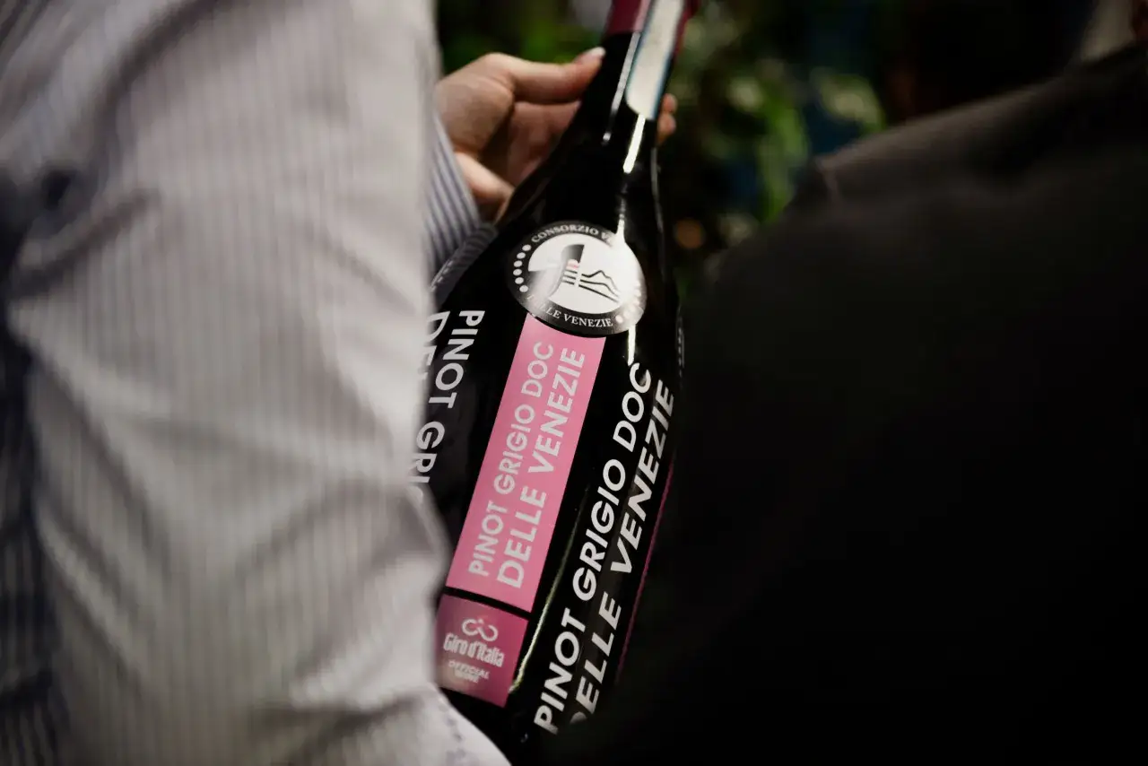 Una mano tiene una bottiglia di Pinot Grigio DOC Delle Venezie, con etichetta rosa e logo Giro d'Italia. Fermo o frizzante?
