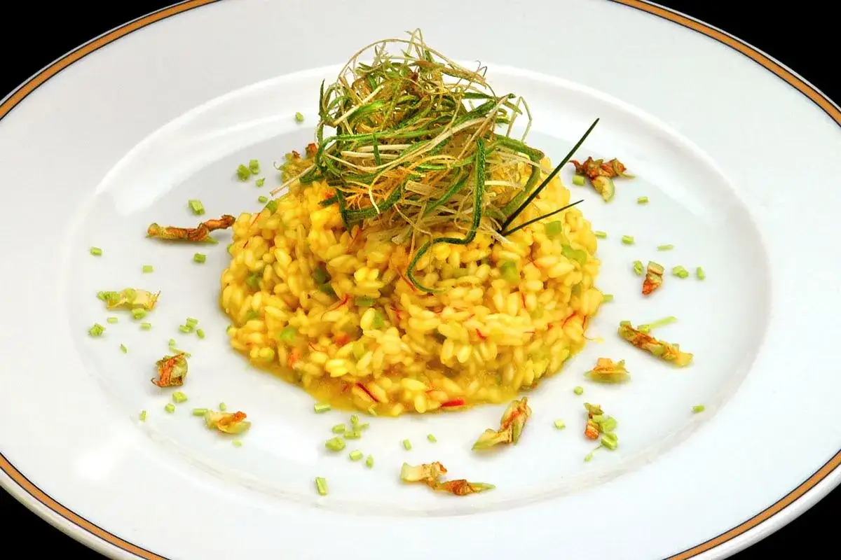 Risotto giallo intenso con fiori di zucca e zafferano, guarnito con erbe croccanti e petali.