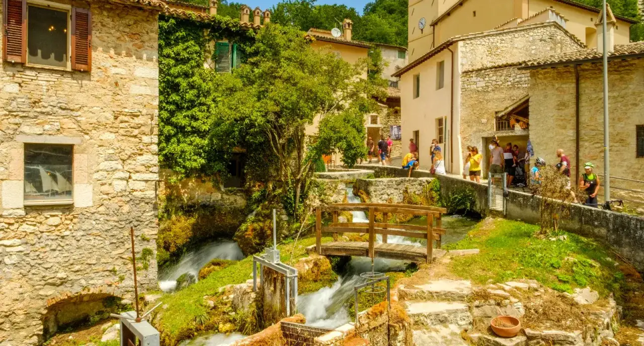 Un borgo incantevole in Umbria dove andare: un ruscello attraversa case in pietra, ponticelli in legno e vegetazione rigogliosa.